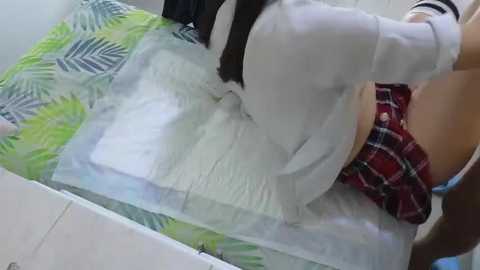 Media: Video of a woman in a white crop top, red plaid skirt, and black leggings, sitting on a bed with green and white leaf-patterned sheets.