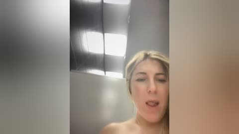 Media: A blurry video of a blonde woman with fair skin, possibly nude, inside a metallic, industrial-looking room with a white ceiling and fluorescent lights.