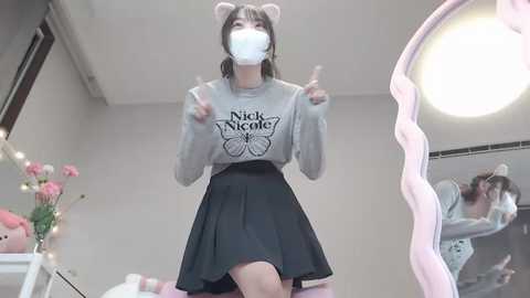 Media: Video of a young woman with a white face mask, gray sweater with \"Nick Viall\" logo, and black skirt, standing in a brightly lit room with pink floral decor and a large mirror.