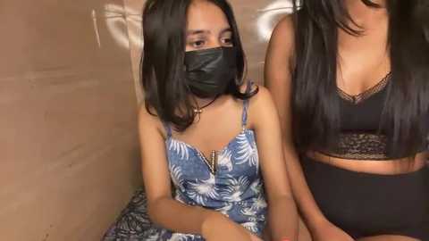 Media: Video of two young women with medium brown skin, long black hair, and masks, sitting on a beige floor. One wears a blue floral dress, the other a black lace bralette.