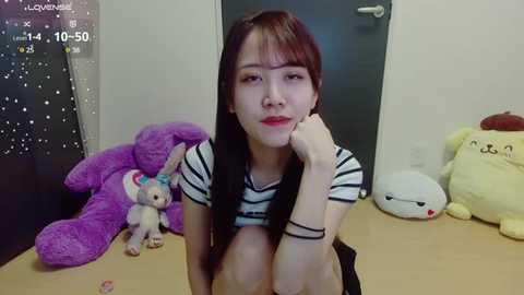 Media: A video of an Asian woman with long brown hair, wearing a striped top, sitting on a wooden table. She has a soft expression. Background includes a door, purple teddy bear, and stuffed toys.