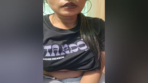 Media: Video of a young woman with medium skin tone, wearing a black t-shirt with \"TARDO\" in bold white letters, and light blue jeans, seated indoors.