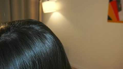 Media: Video of a close-up of a person's back, showing sleek, dark hair styled in a shiny, straight manner. Background features a beige wall with a small framed picture and a soft, warm light source.