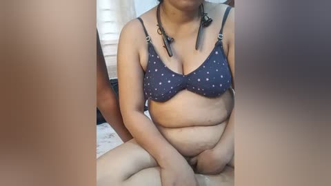 Media: Video of a plus-sized woman with light brown skin, wearing a dark blue bra with white polka dots, sitting on a bed. Background shows a white curtain and a partially visible person's leg.