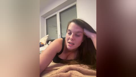 Media: Video of a young woman with light skin, long brown hair, and a black tank top, lying on a bed, looking tired and resting her head on her hand.