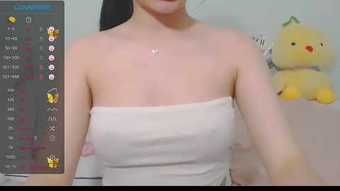 Media: Video of a young woman with fair skin, wearing a white strapless dress, with a small mole on her neck. Background includes a stuffed yellow duck and a light-colored wall.