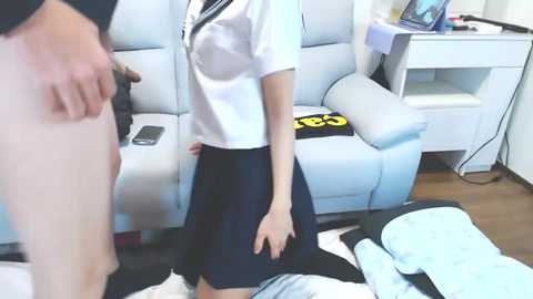 Media: Video of a woman in a white blouse and navy skirt standing in a living room with white furniture, a black purse, and a gray sofa.