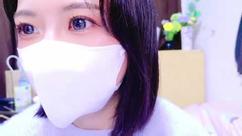 Media: Video of a young Asian woman with fair skin, blue eyes, and straight black hair, wearing a white surgical mask. She is indoors, with a blurred background showing a vase of colorful flowers and a wooden shelf.