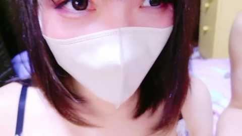 Media: Video of an East Asian woman with long, straight dark hair, wearing a white surgical mask, a black spaghetti strap top, and pale skin. The background is blurred, featuring soft pastel colors and a bed with white linens.