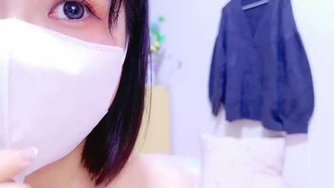 Media: Video of an Asian woman with long black hair, wearing a white face mask, taken from a close-up angle, showing her eye. Background includes a blue jacket hanging on a wall and a blurred room with white and beige furniture.