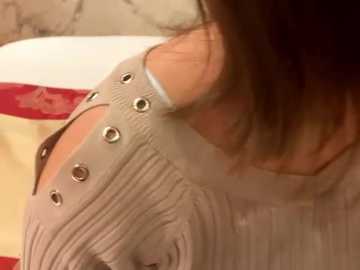 Media: Video of a woman's upper body, wearing a light grey, ribbed sweater with metal eyelet detailing around the neckline, and a white collar. Background features a red and white patterned fabric.