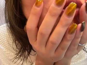 Media: A close-up video of a woman with fair skin, long brown hair, and manicured nails painted in a gradient of golden yellow to metallic gold, covering her mouth. She wears a textured white sweater.