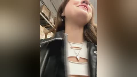 Media: Video of a young woman with fair skin and brown hair, wearing a white crop top and black leather jacket, smiling. Background shows a blurred, beige interior.