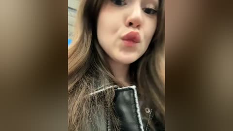 Media: Video of a young woman with long, dark brown hair, fair skin, and light makeup. She's wearing a black leather jacket, puckering her lips, and the background is blurred.