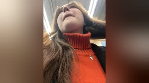 Media: Video of a woman with long brown hair, wearing an orange turtleneck sweater, leaning back with hands on head, under fluorescent lights.