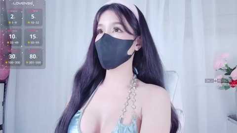 Media: Video of an East Asian woman with long, dark hair, wearing a black face mask, light blue halter top, and silver chain necklace. Background features a pink flower arrangement and a calendar overlay.