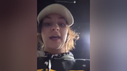 Media: Video of a young woman with fair skin, light brown hair, and a white baseball cap, wearing a black jacket with yellow accents, indoors under dim lighting.