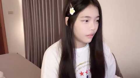 Media: Video of an Asian girl with long black hair, wearing a white T-shirt, sitting indoors next to a window with vertical blinds. She has a white flower hair clip.