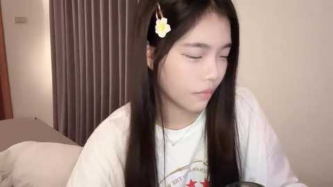 Media: Video of a young Asian woman with long black hair adorned with a yellow flower hairpin, wearing a white T-shirt, sitting on a bed in a dimly lit room with grey curtains in the background.