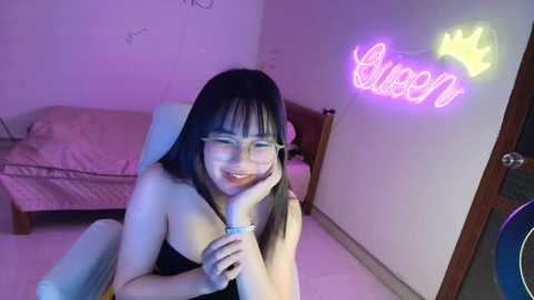 Media: A video of a smiling, fair-skinned woman with long black hair, wearing glasses and a black strapless top, in a bedroom with a pink neon crown sign and a bed.