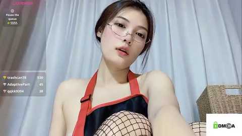 Media: A young Asian woman with fair skin and brown hair, wearing glasses, a black halter top with red straps, and fishnet stockings, poses against a white curtain background.