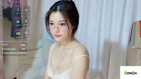 Media: Video of an East Asian woman with light skin and long dark hair, wearing glasses, a white bra, sitting indoors, with a livestream overlay showing viewer count and chat messages.