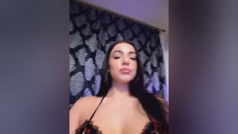 Media: Video of a woman with long black hair, fair skin, wearing a black lace bra, standing against a dark floral-patterned curtain.