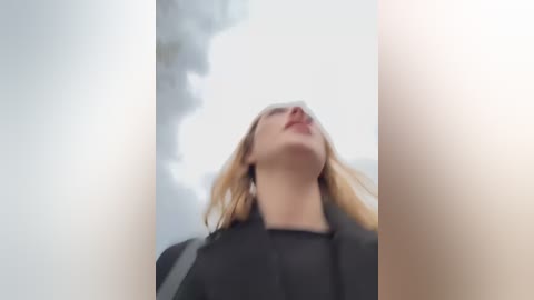 Media: Video of a blonde woman with fair skin, wearing a black coat, looking upward with a serene expression, surrounded by soft, blurred greenery in the background.