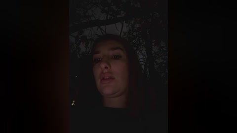 Media: A video of a young woman with light skin, dark hair, and a neutral expression, standing in a dimly lit forest at night. She wears a dark top and is partially obscured by foliage.