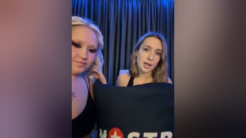 Media: Video of two women with light skin and blonde hair. One wears a black bra, the other a black top, both with makeup. They hold a black Marvel Avengers pillow with a red circle.