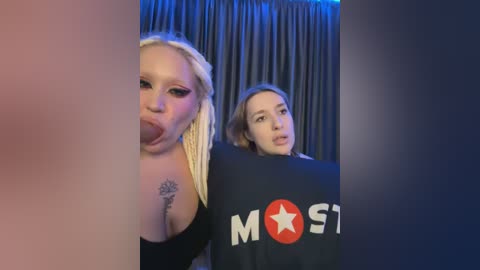 Media: Video of two women with blonde hair and medium builds, one with a star tattoo and a black \"MOST\" shirt, the other in a black top, against dark curtains.