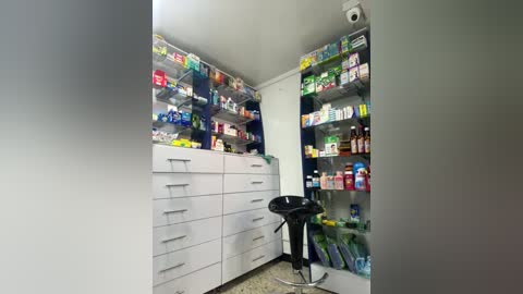 Media: Video of a modern, well-lit pharmacy interior. White cabinets and shelves filled with various medications, vitamins, and cosmetics. Black swivel stool in foreground.