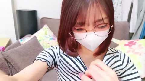 Media: Video of a young Asian woman with straight, shoulder-length brown hair, wearing a white face mask, round glasses, and a striped top, sitting on a bed with colorful pillows and a floral duvet.