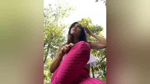 Media: Video of a young woman with medium brown skin and long, straight brown hair, wearing a white top and vibrant pink textured pants, sitting in a lush, green forest.