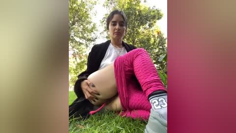 Media: Video of a young woman with medium-dark skin and dark hair, wearing a black jacket, pink leggings, and gray sneakers, sitting on grass outdoors, surrounded by green foliage.