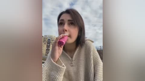 Media: Video of a young woman with straight, shoulder-length brown hair, wearing a beige knit sweater, eating a pink lollipop. Background features a cloudy sky and city buildings.