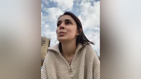 Media: Video of a young woman with light skin and dark hair, wearing a beige knitted hoodie, looking up at a cloudy sky. Background features a tall building and a cityscape.