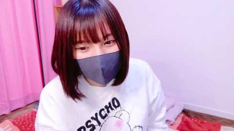 Media: Video of an East Asian woman with straight, shoulder-length brown hair, wearing a white t-shirt with \"Psycho\" and a bunny graphic, a black mask, and sitting on a red and white polka-dot mat. Background features a pink curtain and light purple wall.