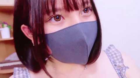 Media: Video of an Asian woman with straight black hair and brown eyes wearing a gray face mask, white checkered top, and earrings. Background includes a blurred wooden shelf with a plant.