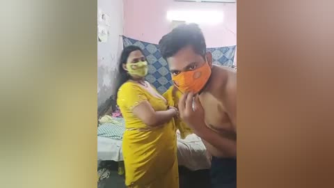 Media: Video of a young man, shirtless, wearing an orange mask, in a dimly lit, cluttered room with a woman in a yellow saree and mask, both looking tense.