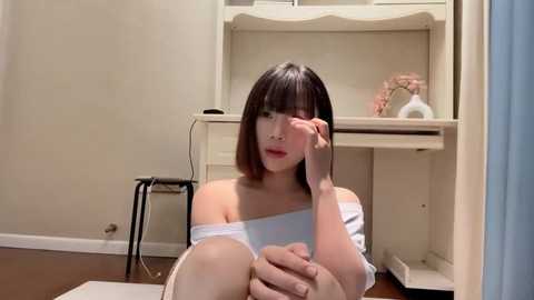 Media: Video of an East Asian woman with shoulder-length black hair, wearing a white off-shoulder top, sitting on the floor, partially covering her face with her hand. Background includes a white desk, a vase, and a black stool.