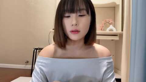 Media: Video of an Asian woman with straight, shoulder-length dark hair, wearing a white off-shoulder top. She has a fair complexion and is seated indoors, with beige walls, a vanity table, and a floral arrangement in the background.