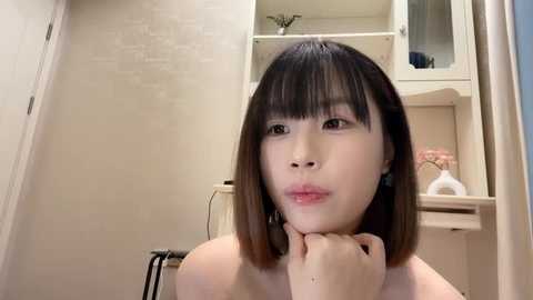 Media: Video of an Asian woman with straight, shoulder-length black hair, light skin, and a contemplative expression, seated in a beige-tiled bathroom with a white shelf and pink flowers in the background.