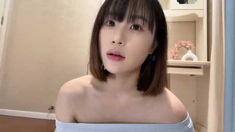 Media: Video of an Asian woman with shoulder-length, straight black hair, wearing a light blue off-shoulder top. She has a neutral expression and is indoors, with beige walls and a white shelf in the background.