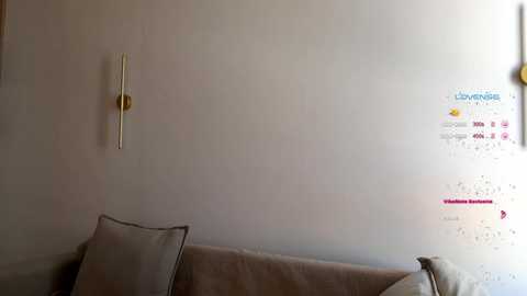 Media: A video of a minimalist, beige-walled room with a modern, gold wall-mounted clock and a cushioned sofa, adorned with a white pillow and decorative cushions.