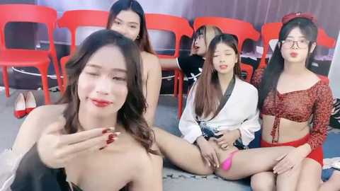 Media: Video of four Asian women in various revealing outfits, sitting on the floor, in a brightly lit room with red chairs in the background.