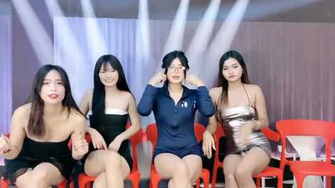 Media: Video of four young Asian women, three in black dresses, one in a blue jumpsuit, seated on red plastic chairs against a white backdrop with white lights.