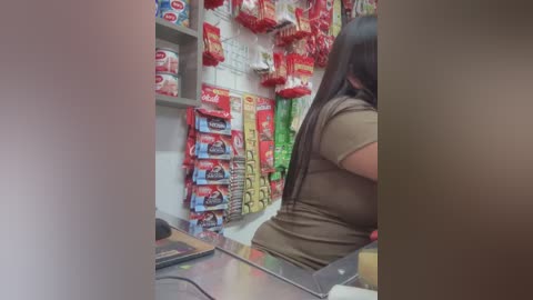 Media: Video of a woman with long hair in a brown top, standing at a snack vending machine in a brightly lit store. Red and green snacks are prominently displayed on the shelves.