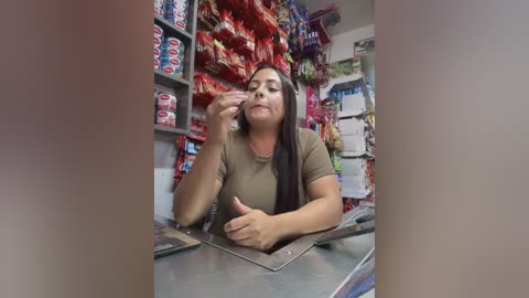 Media: Video of a Latina woman with long black hair, medium build, and olive skin, wearing a brown top, behind a convenience store counter filled with colorful chips and snacks. She's smiling, holding a bag of chips.