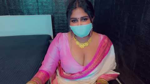 Media: Video of a South Asian woman with dark hair, wearing a pink and white sari, gold necklace, and face mask, sitting on a bed against a dark brick wall.